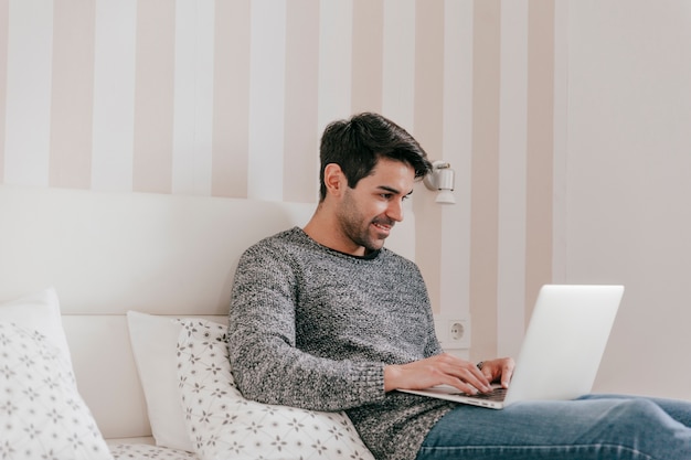 Hombre sonriente con la computadora portátil en la cama