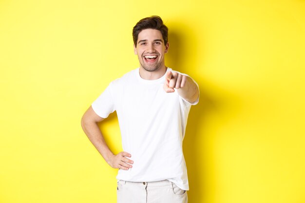 Hombre sonriente confiado apuntando a su cámara, de pie con ropa blanca sobre fondo amarillo.