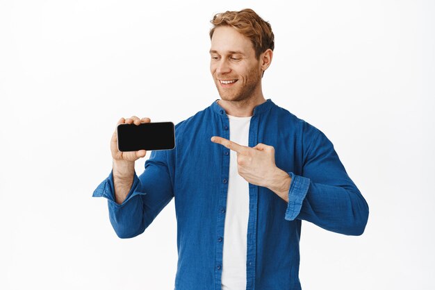 Hombre sonriente confiado señalando y mirando la pantalla horizontal del teléfono inteligente que muestra la aplicación del teléfono móvil con expresión de cara satisfecha de pie sobre fondo blanco