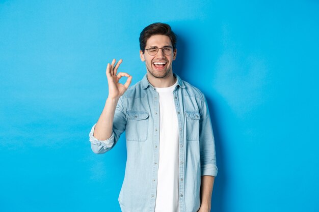 Hombre sonriente confiado en vasos que muestra el signo de ok, guiñando un ojo para garantizar o recomendar algo, fondo azul.