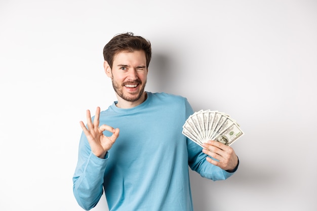 Hombre sonriente descarado guiñando un ojo, mostrando el signo de Ok y sosteniendo dinero, concepto de préstamo o crédito rápido, de pie sobre fondo blanco.