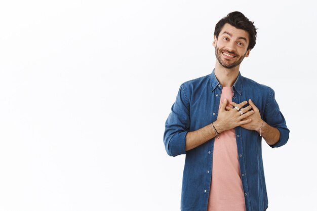 Hombre sonriente encantador y optimista agradeciendo desde el fondo del corazón, presionando las manos contra el pecho y sonriendo lindo mientras expresa gratitud por un regalo genial o agradece la ayuda, de pie, la pared blanca halagada