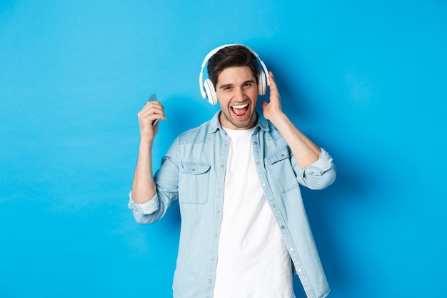 Hombre sonriente feliz disfrutando de escuchar música en auriculares, sosteniendo el teléfono inteligente en la mano levantada, de pie sobre fondo azul.