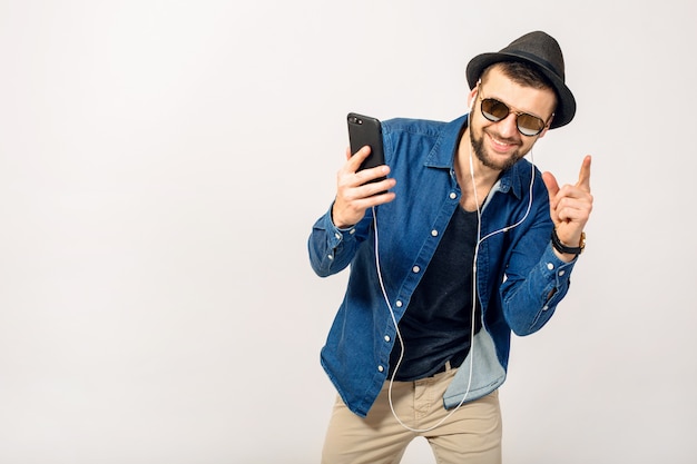 Hombre sonriente feliz guapo joven escuchando música en auriculares aislados sobre fondo blanco de estudio, sosteniendo smartphone, vistiendo camisa vaquera, sombrero y gafas de sol