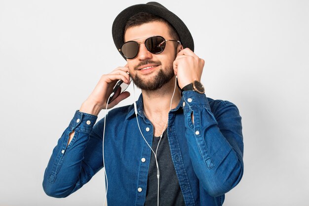 Hombre sonriente feliz guapo joven escuchando música en auriculares aislados sobre fondo blanco de estudio, sosteniendo smartphone, vistiendo camisa vaquera, sombrero y gafas de sol