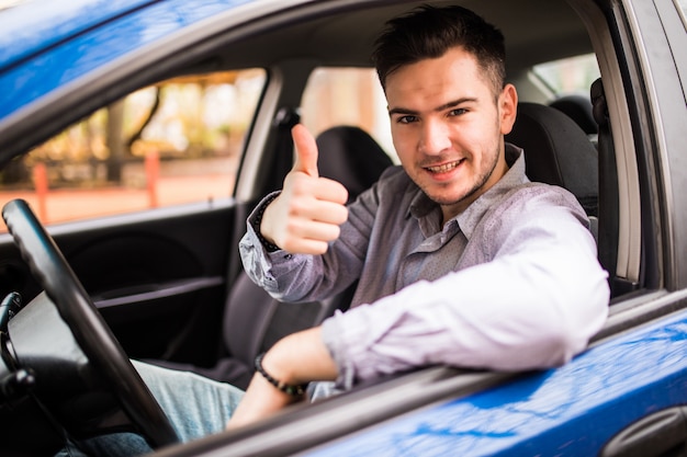 Hombre sonriente feliz que se sienta dentro del coche que muestra los pulgares para arriba. Chico guapo entusiasmado con su nuevo vehículo. Expresión de la cara positiva