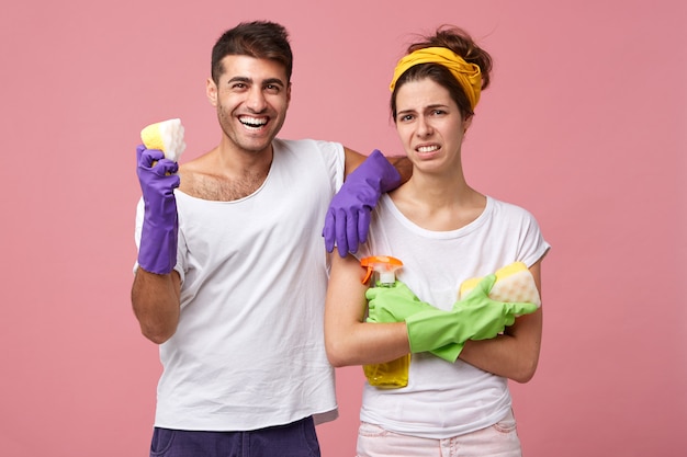 Hombre sonriente con guantes protectores y camiseta blanca sosteniendo una esponja en la mano apoyándose en su bonita esposa que no está satisfecha con no querer hacer la limpieza de pie con las manos cruzadas con el ceño fruncido