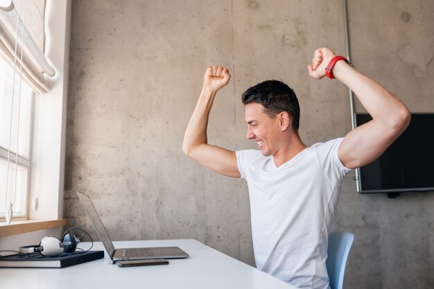 Hombre sonriente guapo joven moderno en traje casual sentado en la mesa trabajando en la computadora portátil, autónomo en casa