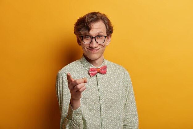 El hombre sonriente guapo positivo le indica, señala con el dedo índice directamente, usa una camisa de moda, elige a alguien, usa gafas, posa sobre una pared amarilla. Deberías unirte a mi