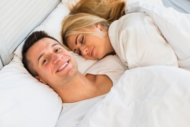 Hombre sonriente joven cerca de mujer que duerme debajo de la manta en cama