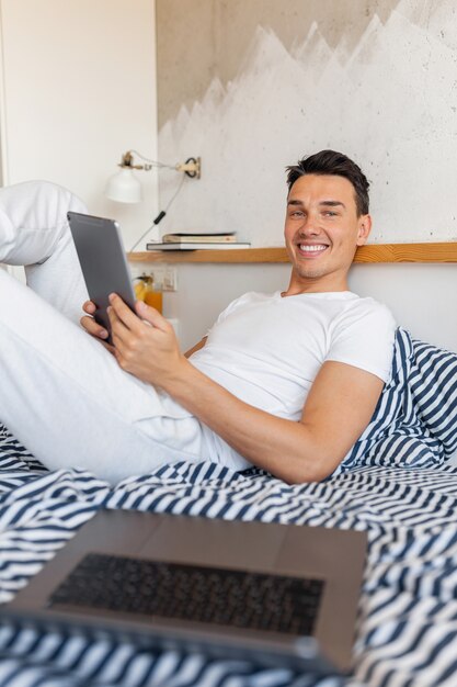 Hombre sonriente joven fresco en traje de pijama casual sentado en la cama en la mañana con tableta, autónomo en casa