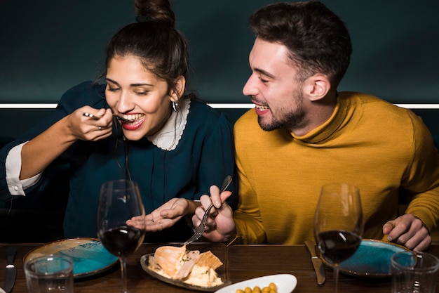 Hombre sonriente joven que mira a la mujer con las bifurcaciones que prueban el queso en la tabla en restaurante