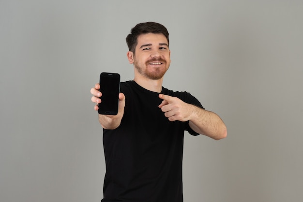 Hombre sonriente joven que muestra su teléfono a la cámara en un gris.