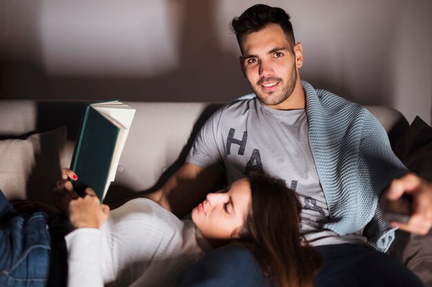 Hombre sonriente joven con el telecontrol y la mujer de la TV con el libro en el sofá