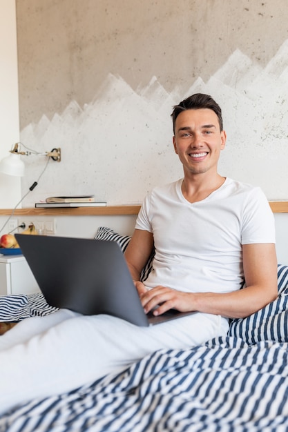 Hombre sonriente joven en traje de pijama casual sentado en la cama en la mañana trabajando en la computadora portátil, autónomo en casa