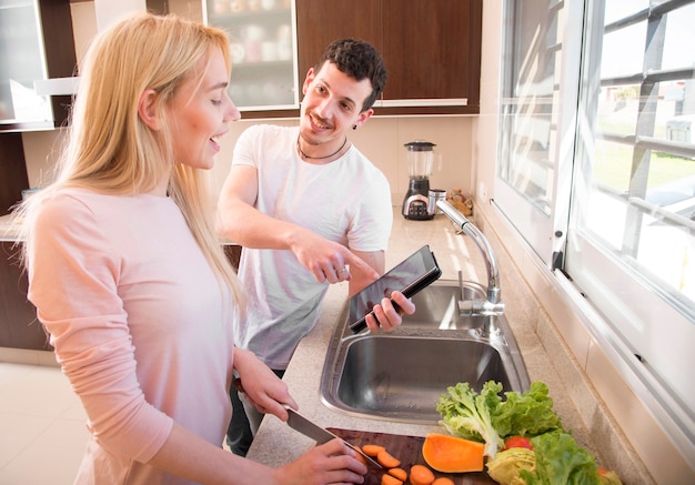 Hombre sonriente mostrando tableta digital a su esposa cortando zanahoria