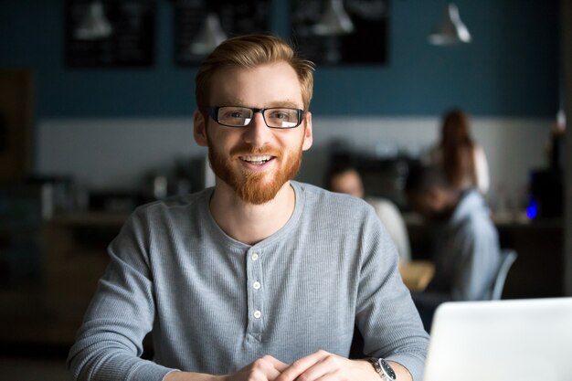Hombre sonriente del pelirrojo con el ordenador portátil que mira la cámara en café