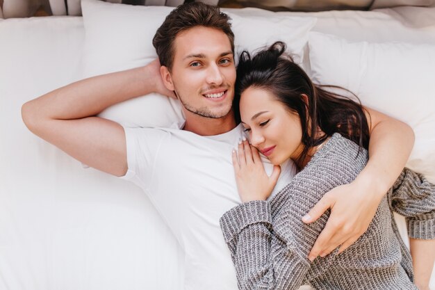Hombre sonriente posando en la cama con esposa durmiendo sobre su pecho
