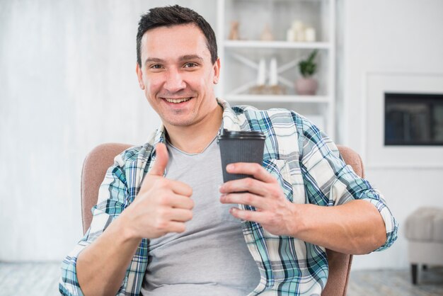 Hombre sonriente con el pulgar hacia arriba sosteniendo la taza de bebida en silla en casa