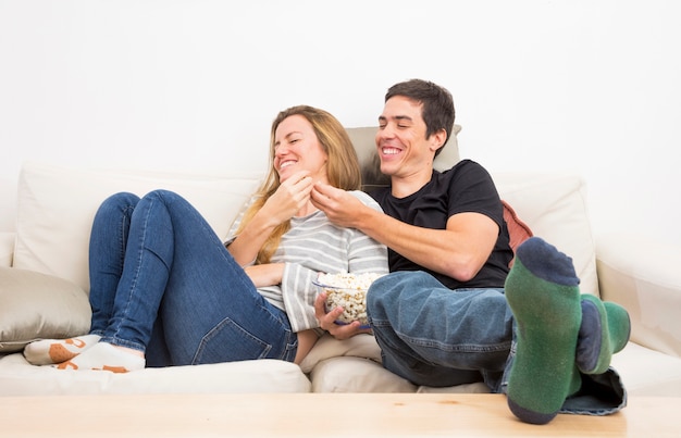 Hombre sonriente que alimenta palomitas a su novia que se sienta en el sofá