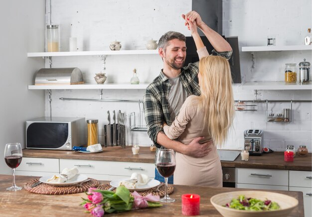 Hombre sonriente que baila con la mujer rubia cerca de la mesa en la cocina