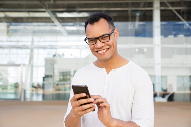Hombre sonriente que se coloca en el edificio de oficinas, sosteniendo el teléfono en manos