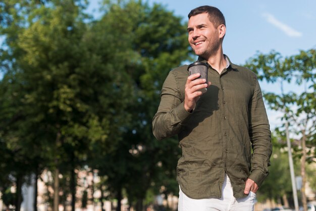 Hombre sonriente que se coloca en el parque que sostiene la taza de café disponible