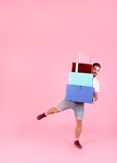 Hombre sonriente que equilibra con la pila de cajas de regalo coloridas contra fondo rosado