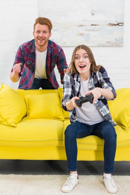 Hombre sonriente que está parado detrás de la mujer emocionada que juega el videojuego en la sala de estar