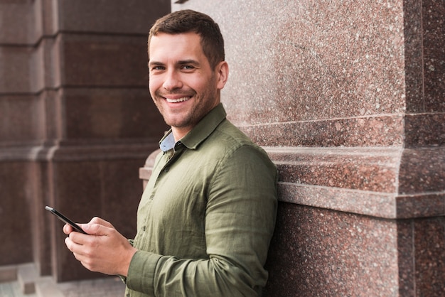 Foto gratuita hombre sonriente que se inclina en la pared que sostiene el teléfono celular y que mira la cámara