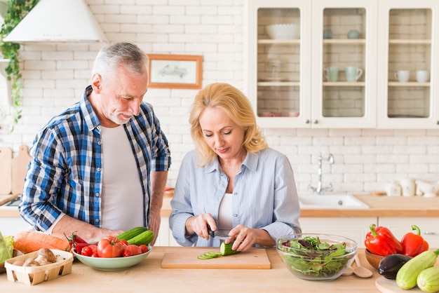 Hombre sonriente que mira a su esposa que corta el pepino con el cuchillo en la tabla en la cocina