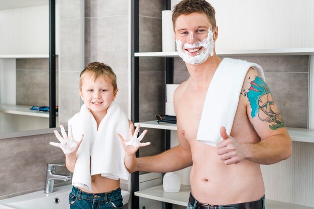 Hombre sonriente que muestra el pulgar encima de la muestra que se coloca con su muchacho que muestra la mano con afeitar espuma en el cuarto de baño