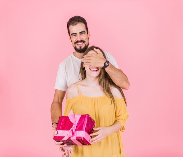 Hombre sonriente que oculta el ojo que da el regalo a su novia sobre fondo rosado