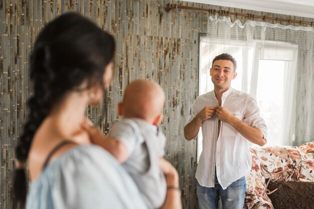 Hombre sonriente que pone en una camisa que mira a su esposa que lleva al bebé en casa