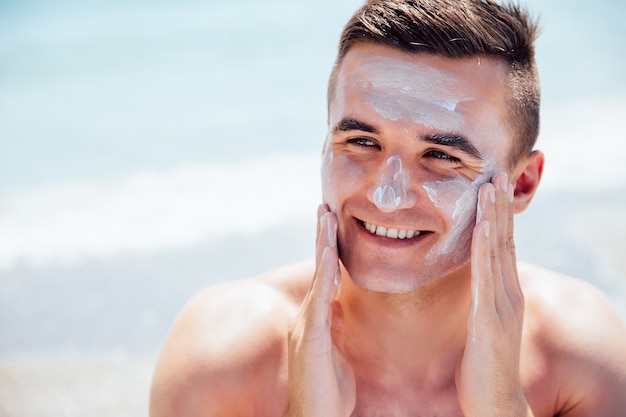 El hombre sonriente que pone la crema que broncea en su cara, toma un sunbath en la playa.