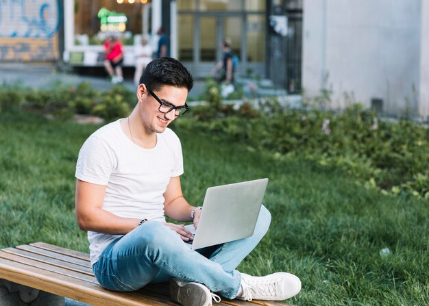 Hombre sonriente que se sienta en el banco que trabaja en la computadora portátil