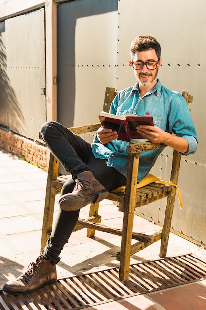 Hombre sonriente que se sienta en la silla de madera que lee el libro