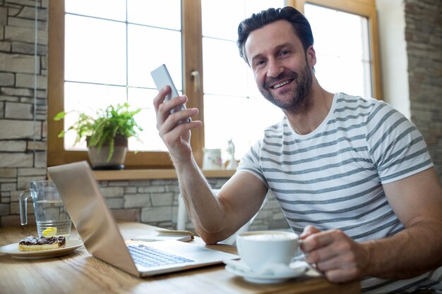 Hombre sonriente que sostiene el teléfono móvil y la taza de café en la cafetería