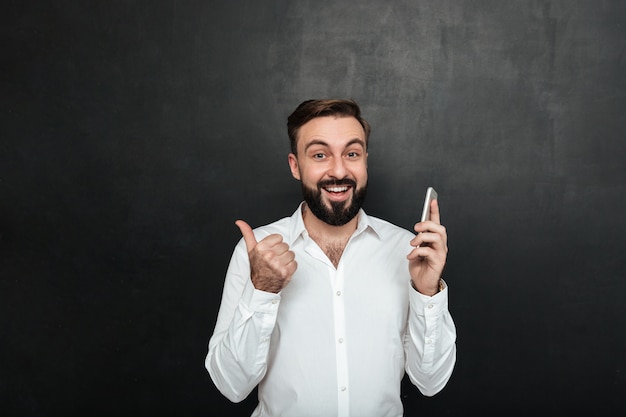 Hombre sonriente satisfecho en camisa blanca con teléfono inteligente y gesticulando con el pulgar a un lado sobre gris oscuro