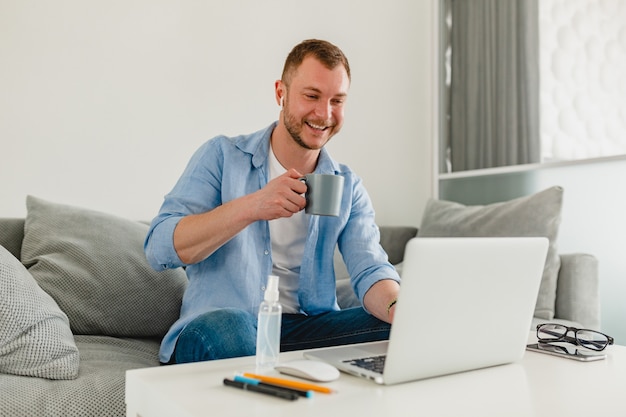Hombre sonriente sentado en el sofá bebiendo té en casa en la mesa trabajando en línea en la computadora portátil