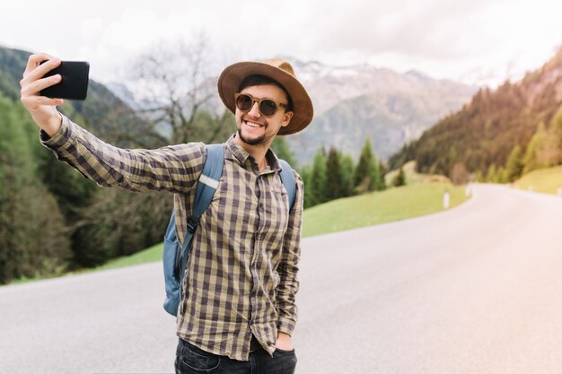 Hombre sonriente con sombrero marrón de pie con la mano en el bolsillo y haciendo selfie mientras atrapa el coche en la carretera