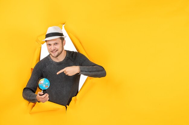 Foto gratuita hombre sonriente con un sombrero sosteniendo un pequeño globo y apuntando un pequeño globo en un rasgado en la pared amarilla