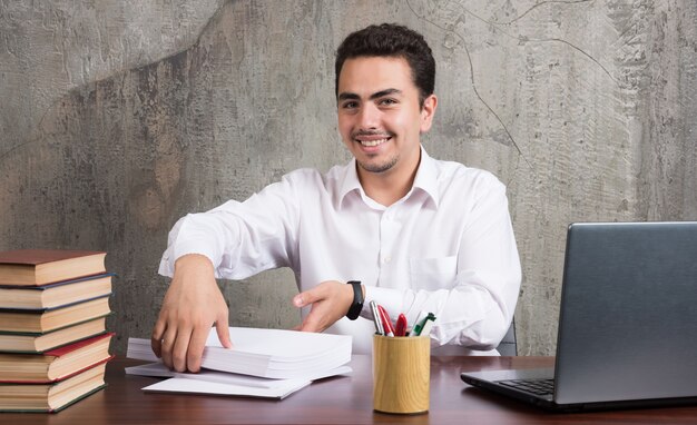 Hombre sonriente sosteniendo hojas de papel y sentado en el escritorio. Foto de alta calidad