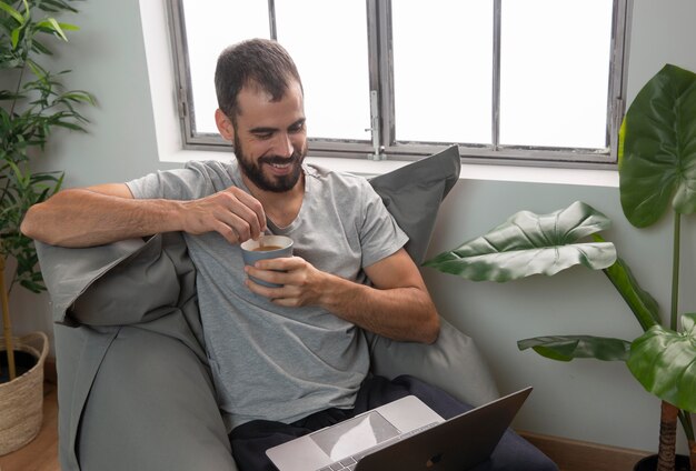 Hombre sonriente tomando café mientras trabaja desde casa en la computadora portátil