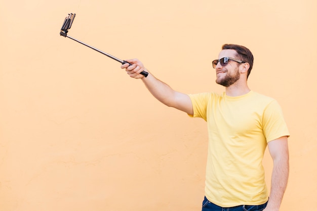 Foto gratuita hombre sonriente tomando selfie en teléfono celular de pie cerca de la pared de durazno