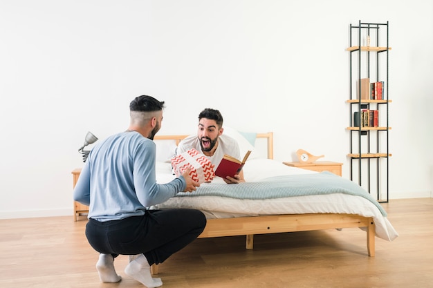 Foto gratuita hombre sorprendido acostado en la cama sosteniendo un libro en la mano mirando a su novio dando caja de regalo