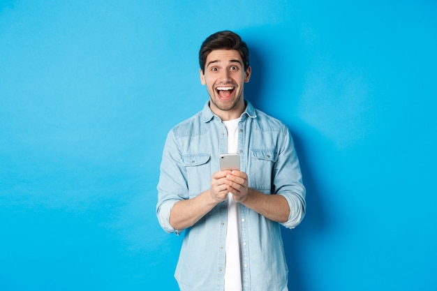 Hombre sorprendido y feliz ganando algo en línea, sosteniendo un teléfono inteligente y regocijándose, de pie contra el fondo azul