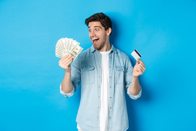 Hombre sorprendido y feliz con tarjeta de crédito, mirando el dinero satisfecho, de pie sobre fondo azul.