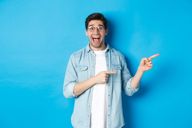 Hombre sorprendido con gafas apuntando a la derecha en el espacio de la copia, mostrando la oferta promocional sobre fondo azul, de pie sobre fondo azul