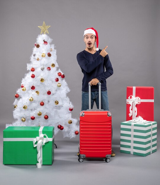 Hombre sorprendido con gorro de Papá Noel cerca del árbol de Navidad blanco con coloridos juguetes de Navidad en